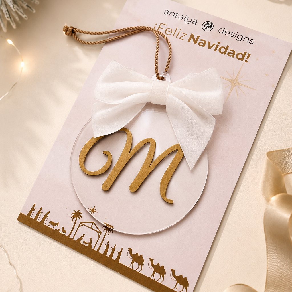 A festive Christmas ornament card from Antalya Designs featuring a gold monogram letter 'M' inside a white oval, tied with a white fabric bow and hanging loop, displayed on a decorated beige holiday background.