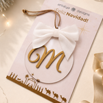 A festive Christmas ornament card from Antalya Designs featuring a gold monogram letter 'M' inside a white oval, tied with a white fabric bow and hanging loop, displayed on a decorated beige holiday background.