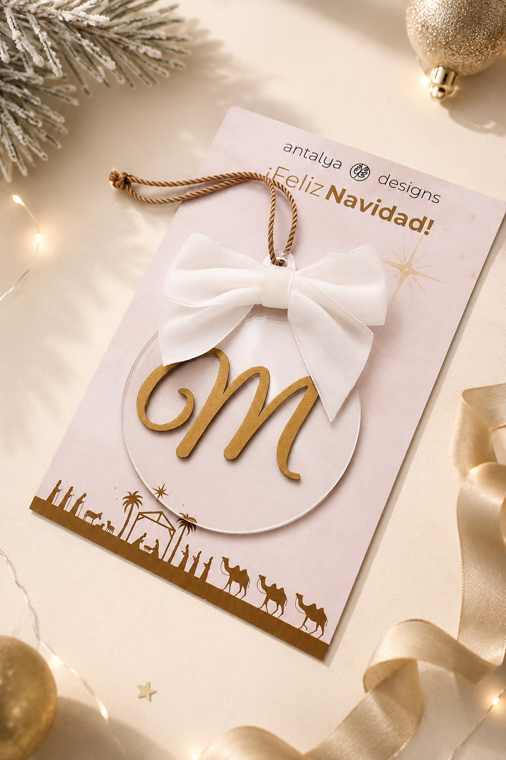 A festive Christmas ornament card from Antalya Designs featuring a gold monogram letter 'M' inside a white oval, tied with a white fabric bow and hanging loop, displayed on a decorated beige holiday background.