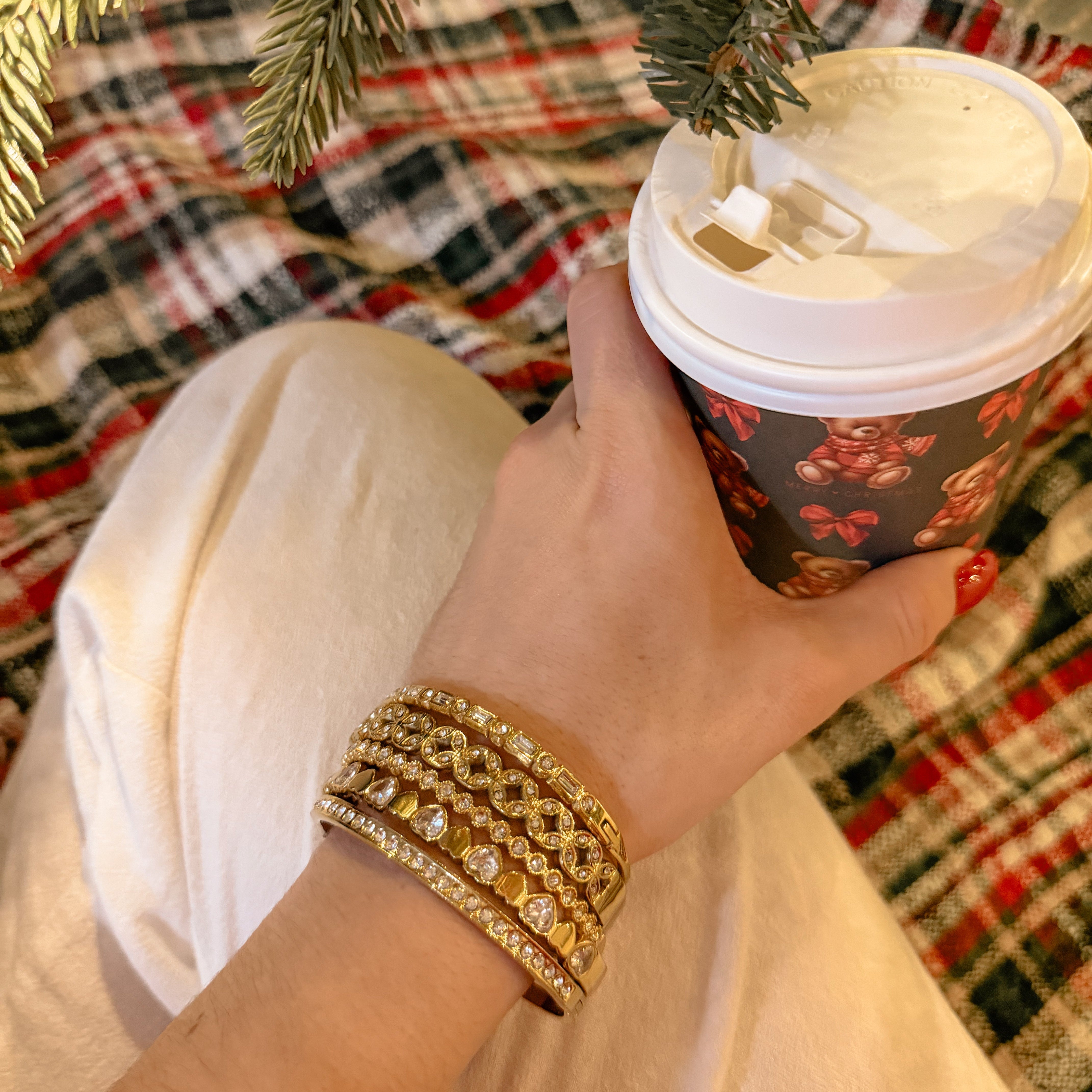 Hand featuring golden bracelets with rhinestones holding a festive coffee cup with a Christmas tree in the background
